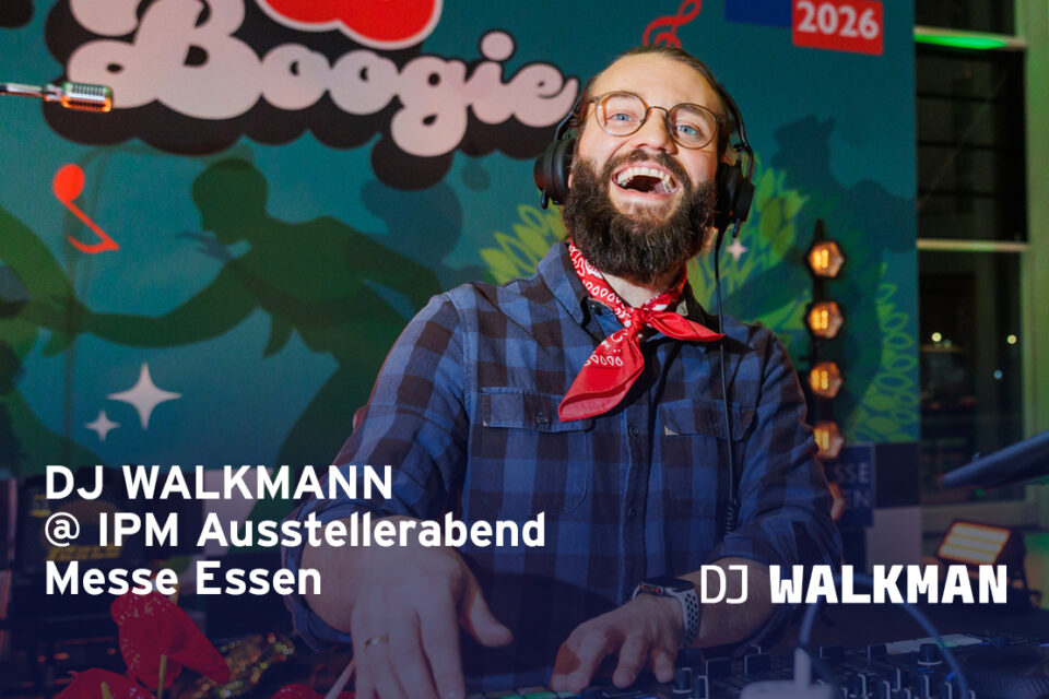 Ein DJ mit Kopfhörern und Bart steht an einem Mischpult und lächelt. Er trägt ein blau kariertes Hemd und ein rotes Halstuch. Aufnahme vom IPM Ausstellerabend in der Messe Essen.