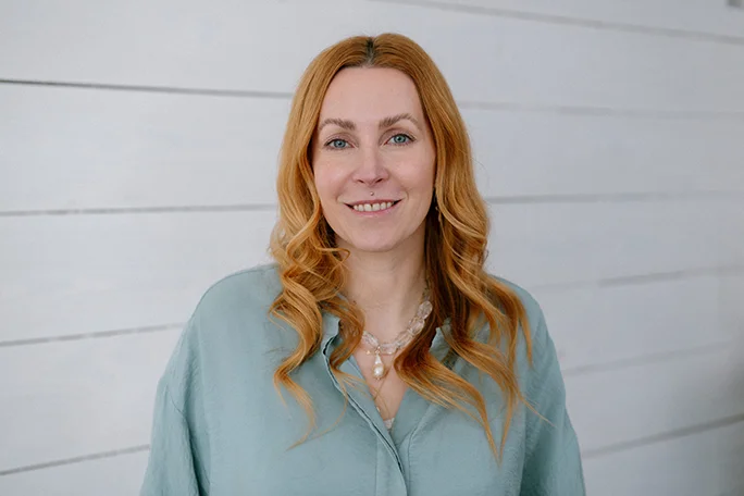 Portrait einer Frau mit kurzen roten Haaren, blauen Augen und Perlenkette vor Holz-Hintergrund.