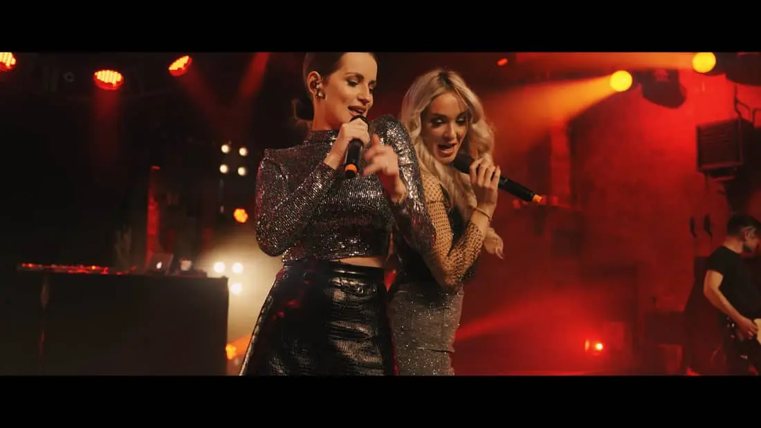 Zwei weibliche Musikerinnen bei energiegeladener Live-Performance mit funkelnden Outfits und stimmungsvoller Bühnenbeleuchtung.