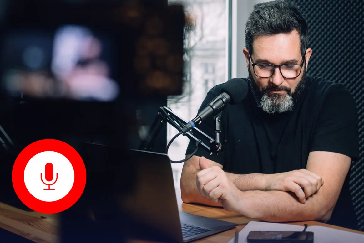 Mann mit Brille und Bart sitzt vor einem Mikrofon und Laptop in einem Tonstudio, während er sich auf eine Audioaufnahme vorbereitet.