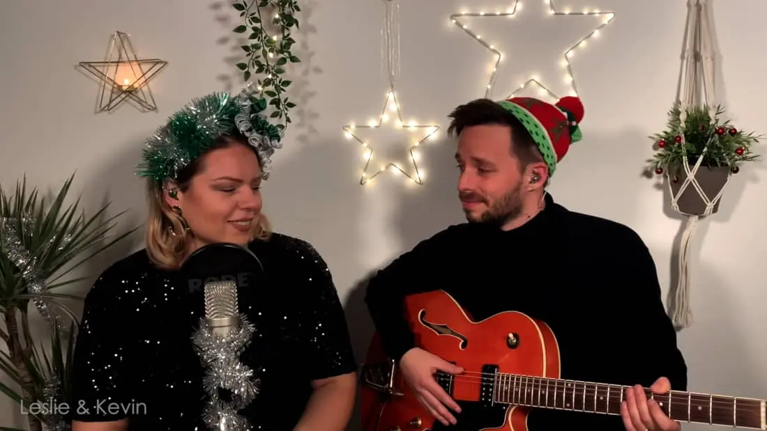 Festliche Musiker Leslie und Kevin bei Weihnachtsperformance mit Gartendekoration und Weihnachtsmützen.
